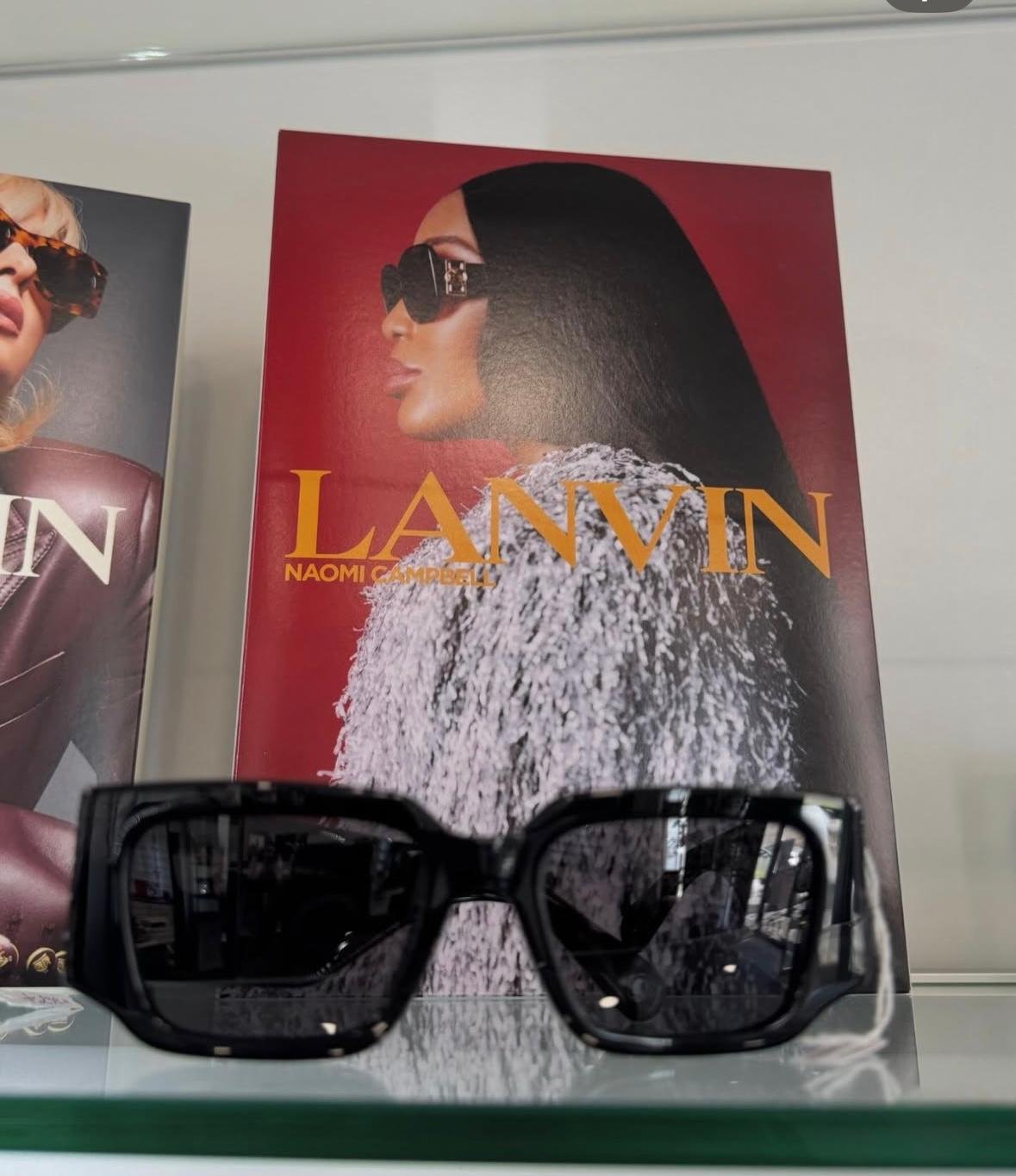 Occhiali da sole neri squadrati su ripiano in vetro davanti a un pannello rosso Lanvin con Naomi Campbell