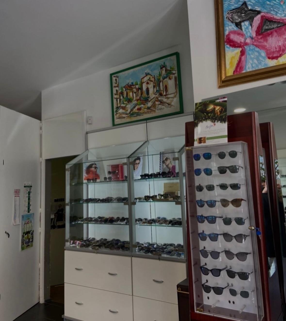 Vetrine in vetro ed espositore in legno con occhiali da sole sotto dipinti colorati di un borgo e un fiocco rosa