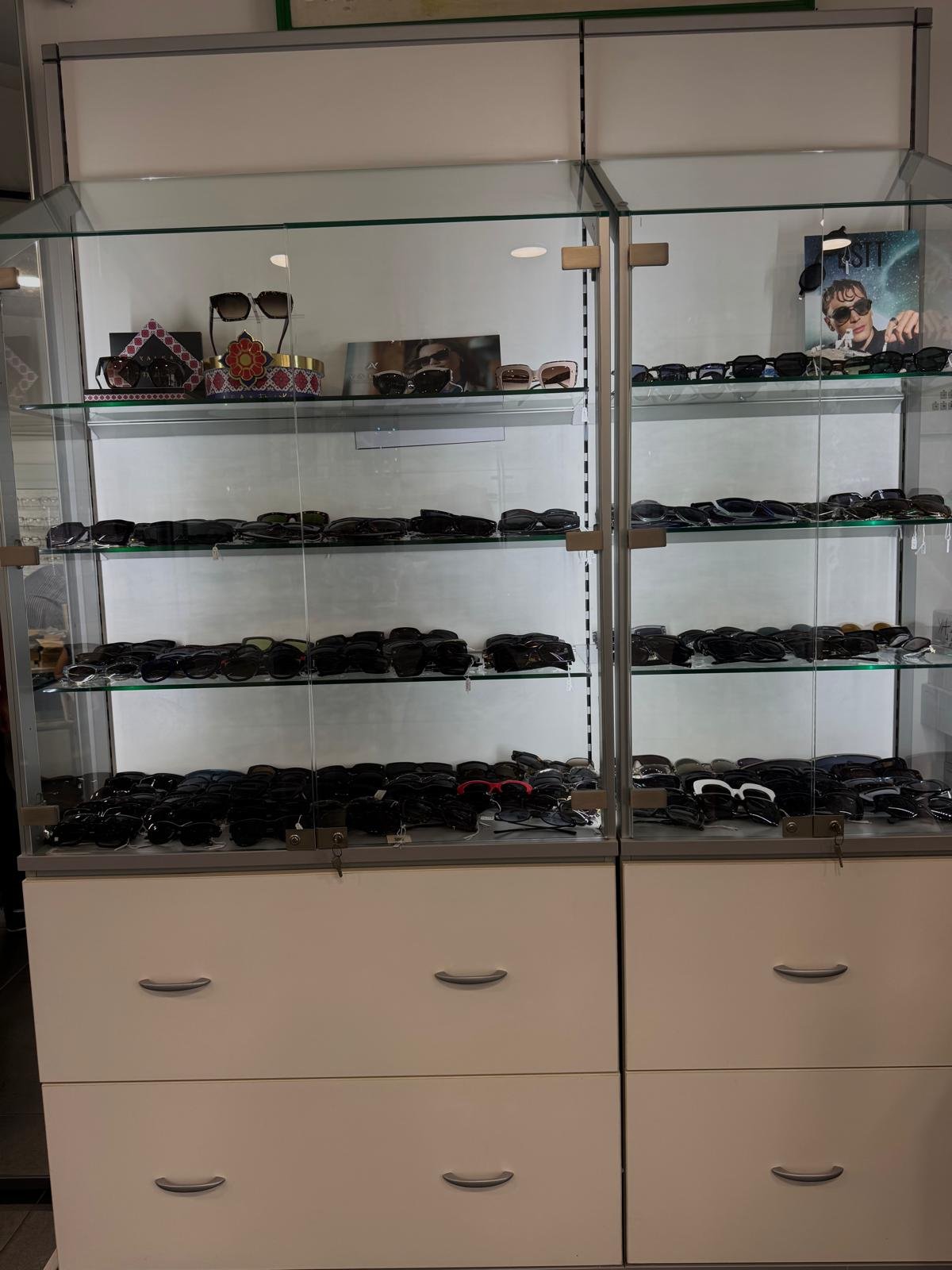 Vetrina in vetro con occhiali da sole di Ottica Medical su ripiani illuminati, sopra cassettiere bianche con maniglie metalliche